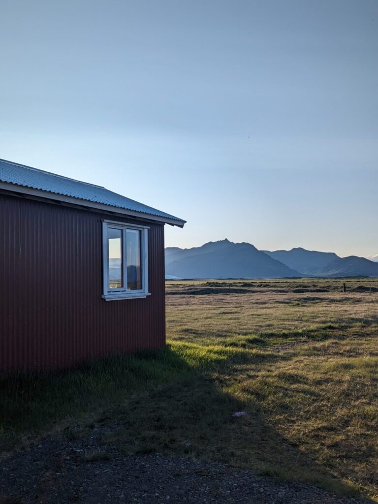 Small red cabin with a red roof sitting in a field with mountain in the distance, The lighting is that of an early sunset