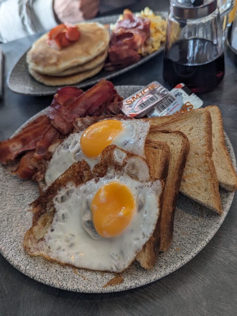 Two plates of food. In the foreground there is a grey plate with two sunny side up eggs, four slices of toast, and three slices of bacon. The background plate has the pancakes, bacon and scrambled eggs.