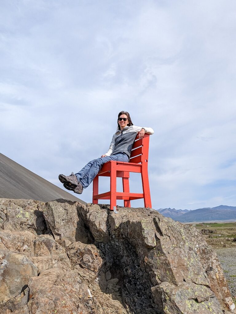 Gabi, a brown haired woman sitting on a large red chair on top of a rock. Gabi is wearing grey hiking boots, blue grey hiking pants, and a grey and white block pull over fleece sweater.