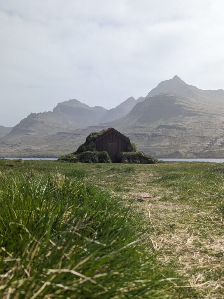 small wooden hut with a grass roof sitting at the edge of a fjord with the mountain in the background.