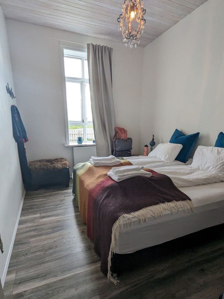 Small rectangular bedroom with two twin beds against the wall on the right. They are covered with a rainbow blanket. There is a window with a tan curtain half open on the far back wall.