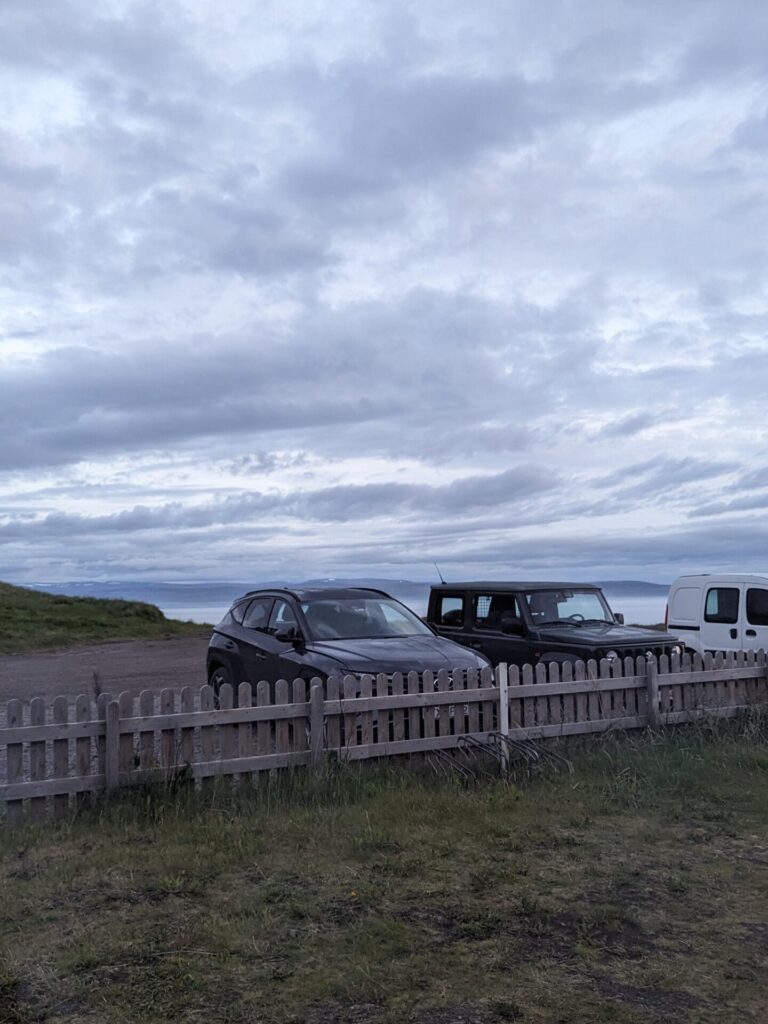 Looking at three cars parked behind a brown wooden fence. The sky is grey and cloudy, the ocean is in the background. 