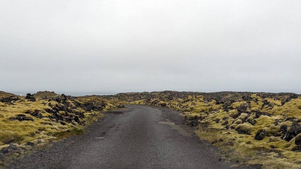 Narrow 1 lane road. Paved but not well maintained. The road is surrounded by mossy lava rocks. The sky is grey and cloudy