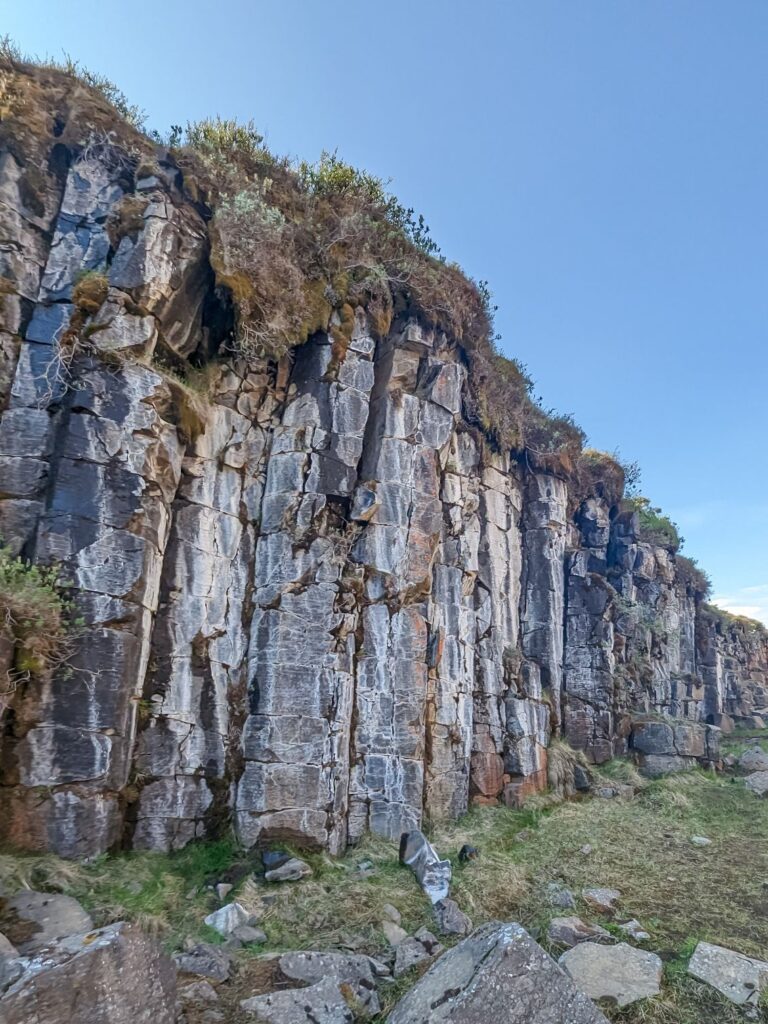 grey cliff with grass covering the top. The cliffside is basalt with white and grey grooves running down it.