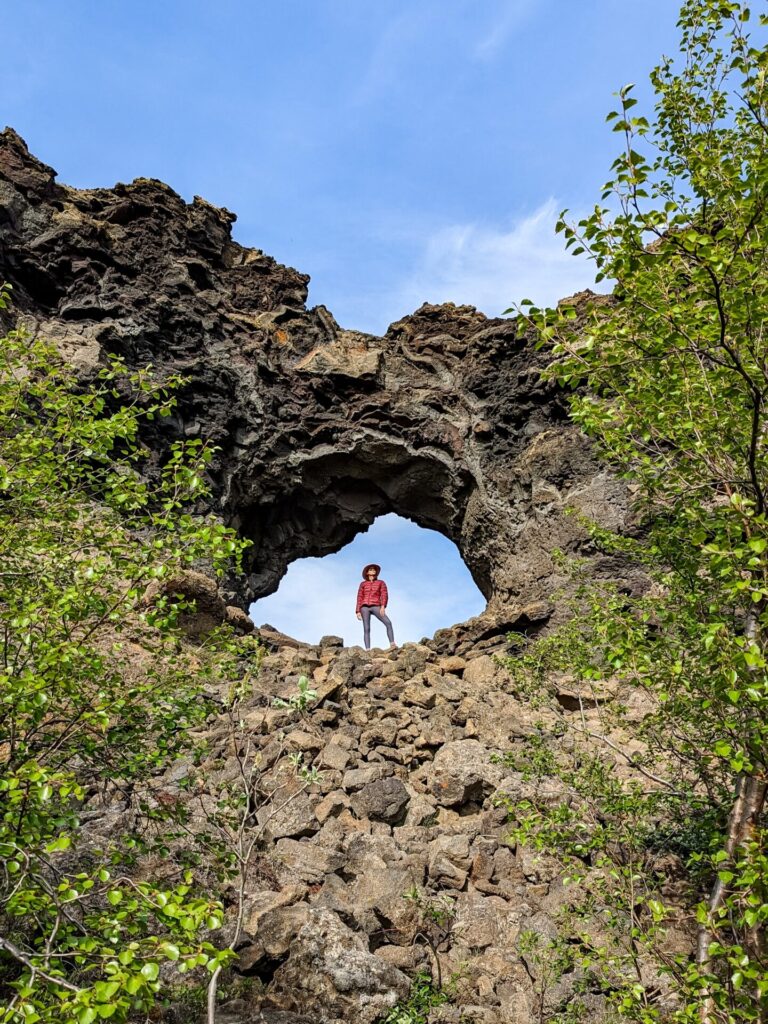 Gabi, a woman in grey leggings, a red puffer jackets, and a wide brim pink hat standing above in the distance inside of a lava rock opening.