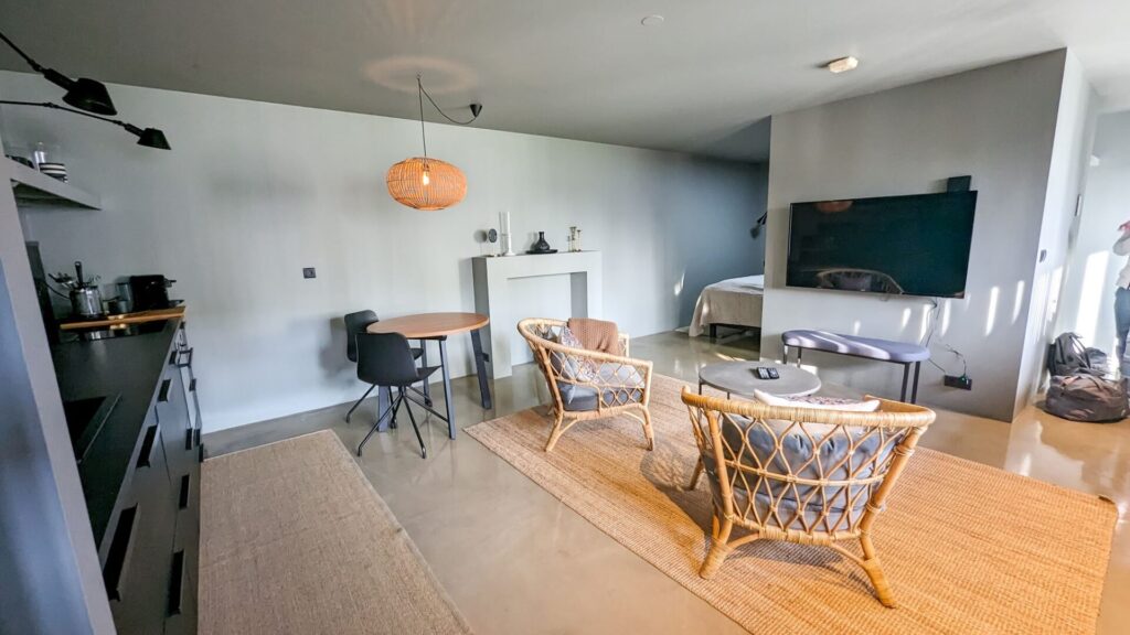 Studio style apartment with a small kitchen, round table, two wicker chairs, a TV, and bed in the background partially concealed behind a wall.