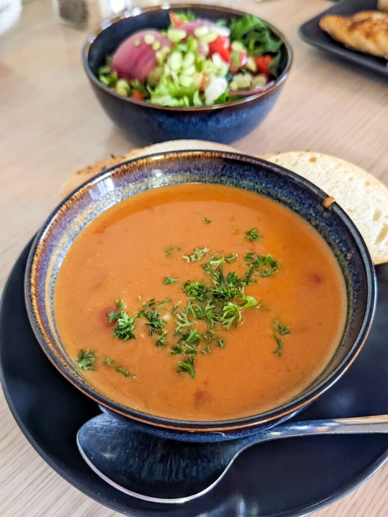 orange red fish soup with parsley on top in a blue bowl. A green and purple salad is in another blue bowl in the background.