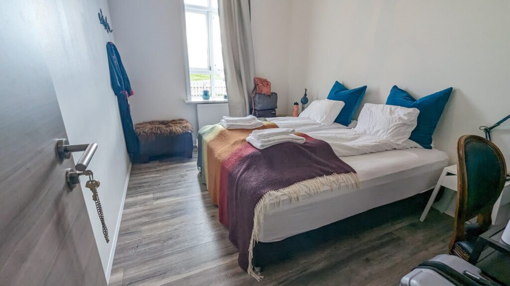 Small rectangular bedroom with two twin beds against the wall on the right. They are covered with a rainbow blanket. There is a window with a tan curtain half open on the far back wall.