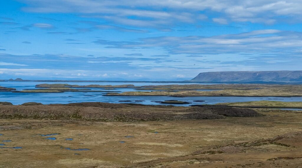 expansive view over a shallow fjord and mossy grassy field with some small hills. In the far background is a mountain on the right side.