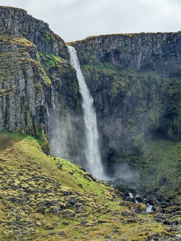 Narrow waterfall cascading over a mossy basalt column cliff into a river.