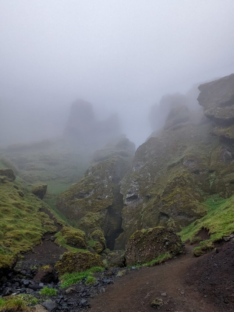 Mossy foggy gorge entrance with the 
dirt trail leading up to it.