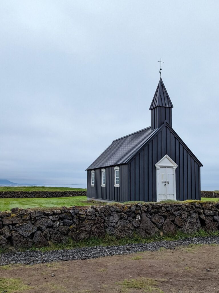 Little black church with white double doors sitting in a grassy field surrounded by a short stone wall.