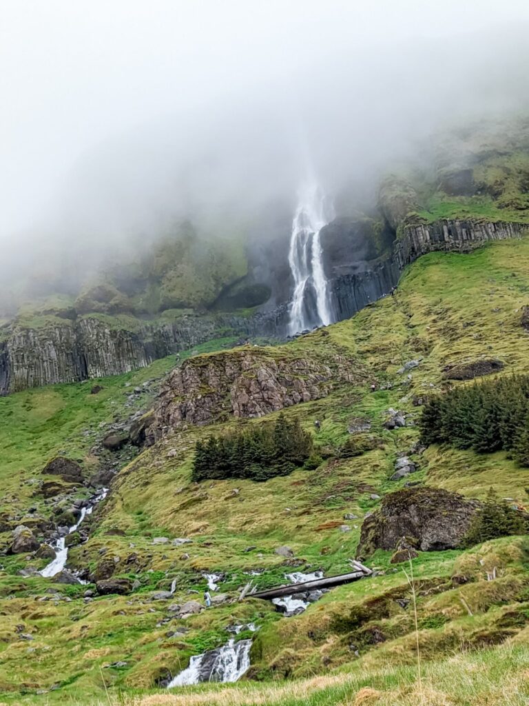 Waterfall cascading over grassy basalt cliffs into a river that runs under a small wooden bridge. The top half of the waterfall is covered in fog.