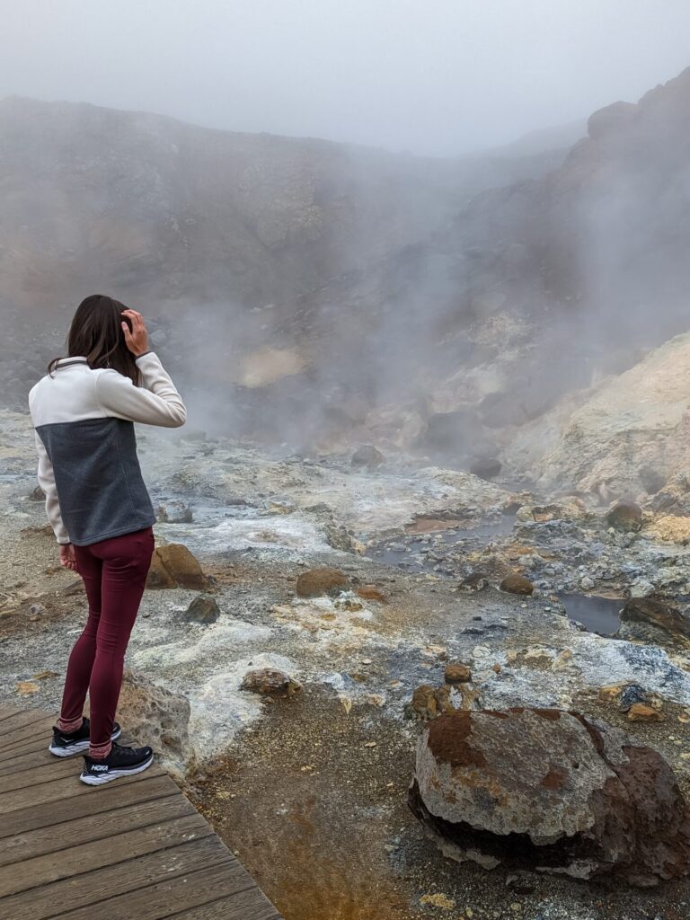 geothermal area with bubbling steaming mud pools. Gabi, a woman with straight brown hair, a color block grey and white fleece pullover and maroon leggings is standing on a wooden walkway looking at the geothermal area.