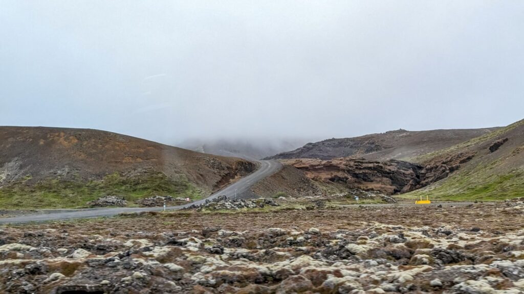 Mossy lava field with a road cresting over a hill in the background. Fog is covering most of the sky and mountains.