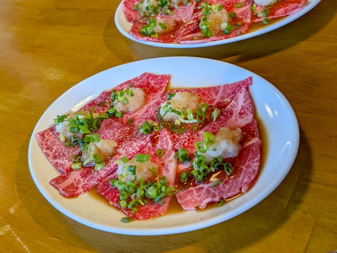 6 sliced of raw wagyu beef sit on white plate. Each piece if topped with a sauce, grated radish, and green onion slices.