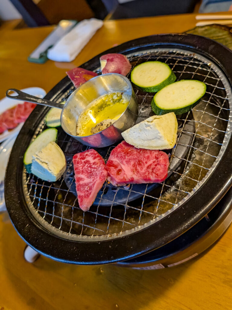 Japanese charcoal grill with a small metal bowl of oil and garlic in the center. Placed around the grill are vegetables and slices of wagyu beef