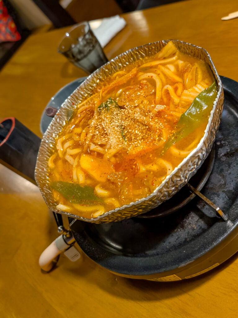 jjigae udon soup. An orange broth soup in a silver metal bowl with udon noodles inside.