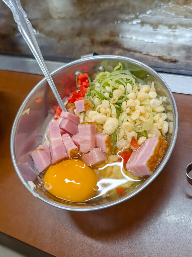 metal bowl filled with ingredients; raw egg, thick sliced bacon, green onions, rice puffs, rice cakes, and cabbage
