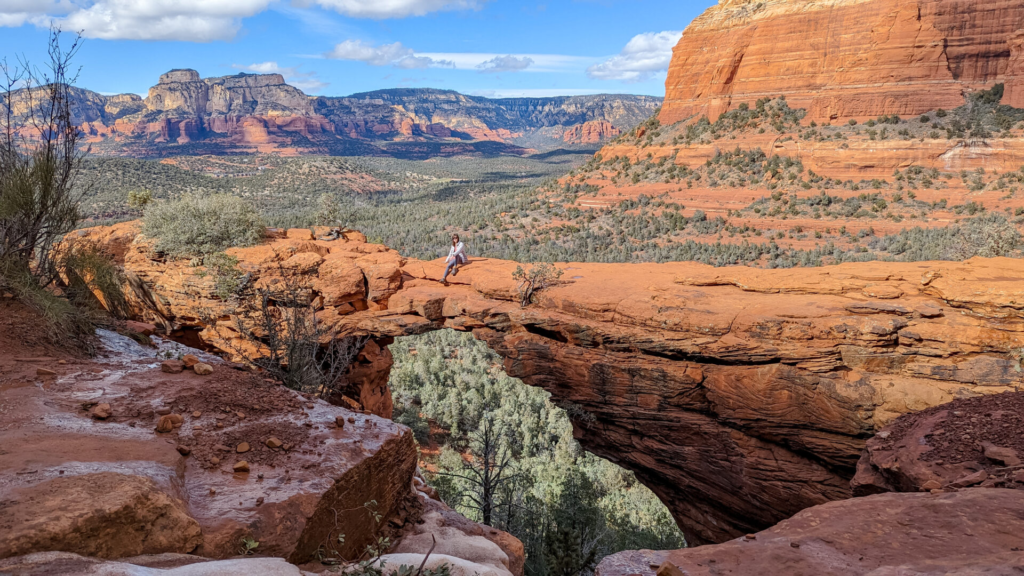 A panoramic landscape shot of a hiker sitting on the middle of the iconic Devil's Bridge sandstone arch. In the background, expansive red rock canyons, green valleys, and layered cliffs stretch out under a bright blue sky filled with puffy white clouds.
