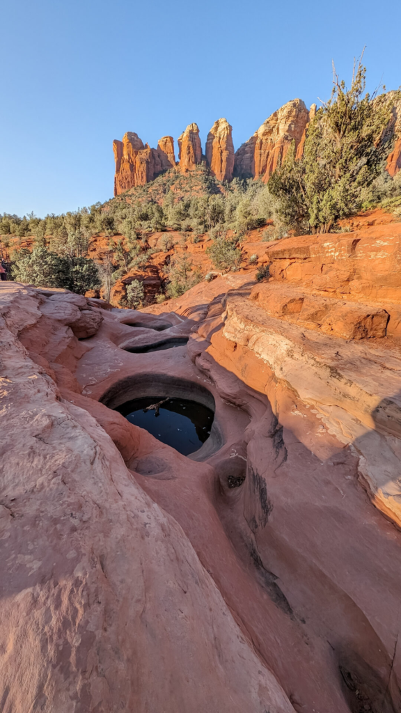A vertical photo showing natural carved rock basins (the Seven Sacred Pools) in reddish-orange sandstone, some holding still water reflecting the sky. Jagged red rock spires towering in the background under a clear sky.