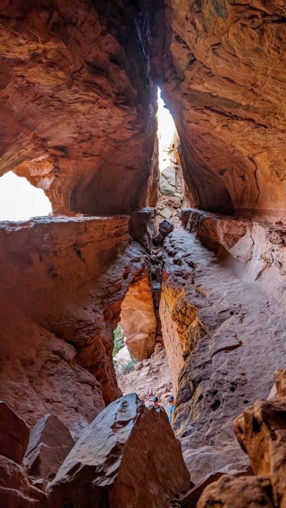 A vertical perspective from inside a deep sandstone cave, looking down through a narrow, jagged rock fissure. Warm daylight filters in from top openings, illuminating layered orange and brown rock walls and hikers visible near the bottom opening.