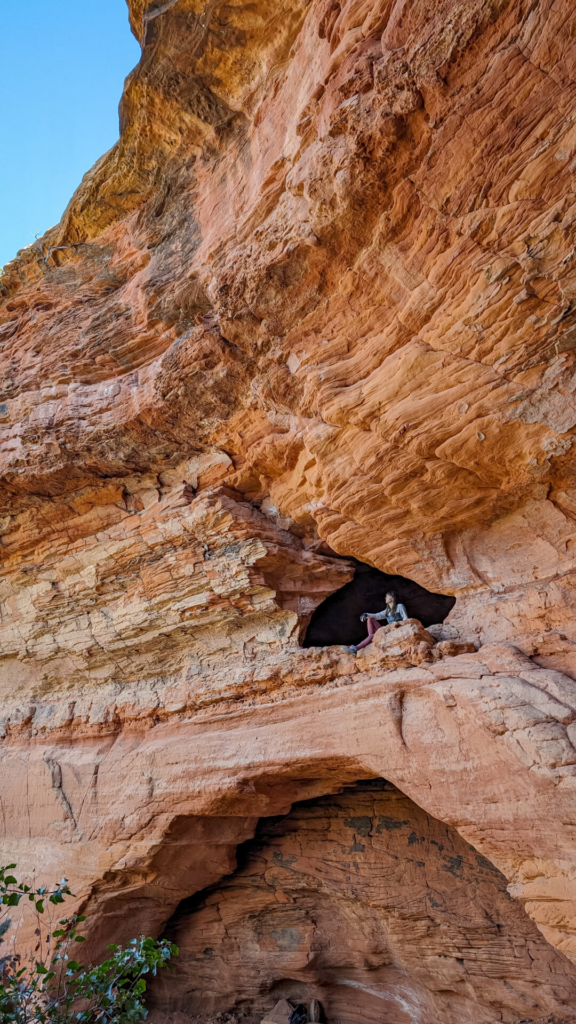 A vertical low-angle shot of a massive, textured orange sandstone cliff face. A lone hiker rests inside a dark, elevated cave alcove naturally hollowed into the middle of the sheer red rock wall.