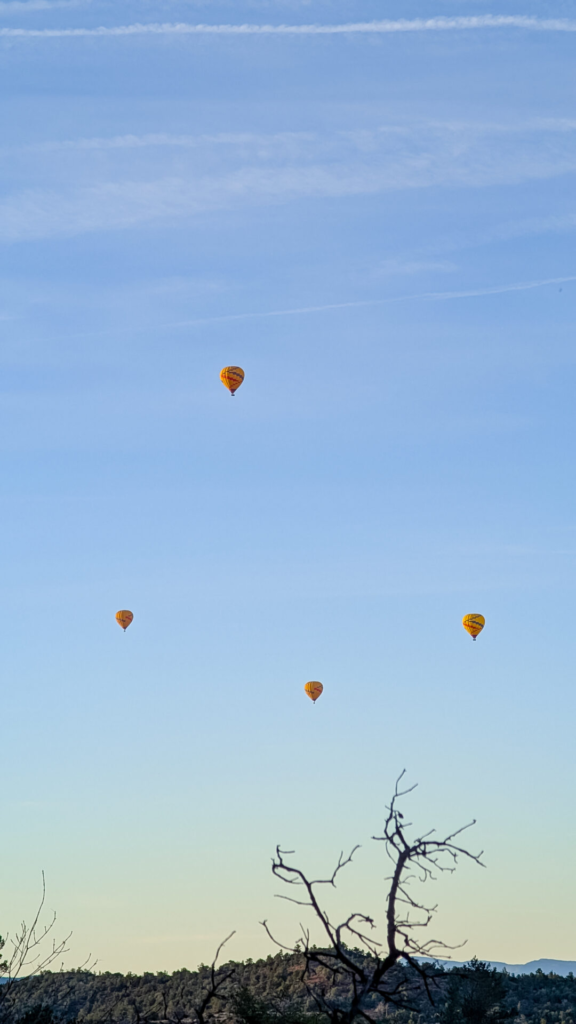 A wide vertical shot of a bright blue morning sky with faint contrails, featuring four yellow hot air balloons floating in formation above a silhouette of tree branches and distant desert hills.