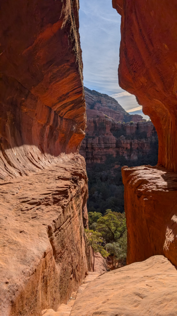 A vertical photo standing on a narrow, sandy red rock ledge within a keyhole-style cave formation. Massive sandstone walls frame both sides, casting warm shadows, and open up to a view of a vast canyon with layered red rock mountains and green treetops under a bright sky.