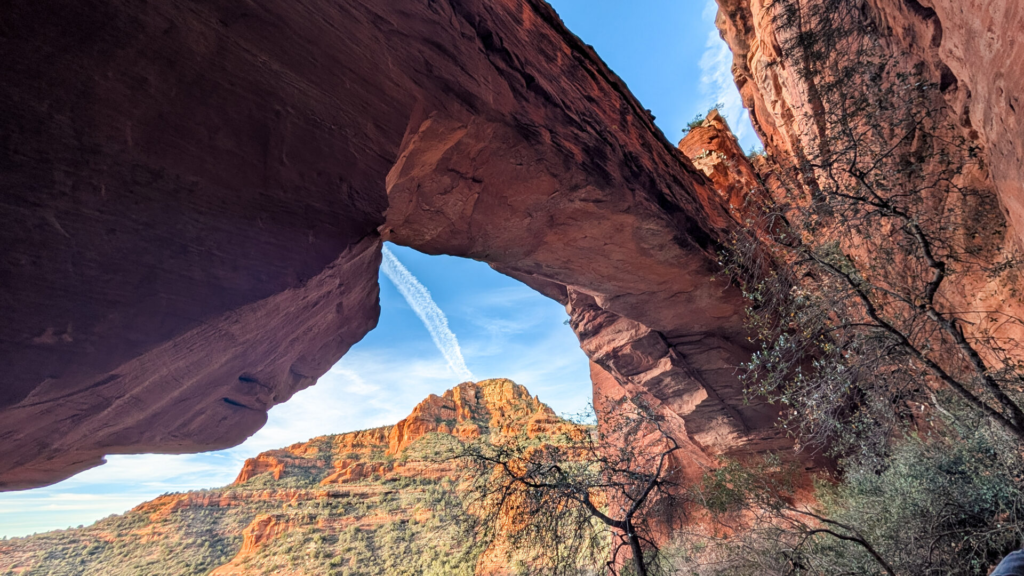 A low-angle shot under a large, shadowy red rock bridge arch. The arch frames a sunlit red rock mountain peak against a clear blue sky with a white jet contrail streak. Sparse trees and green foliage hug the right side of the canyon wall.