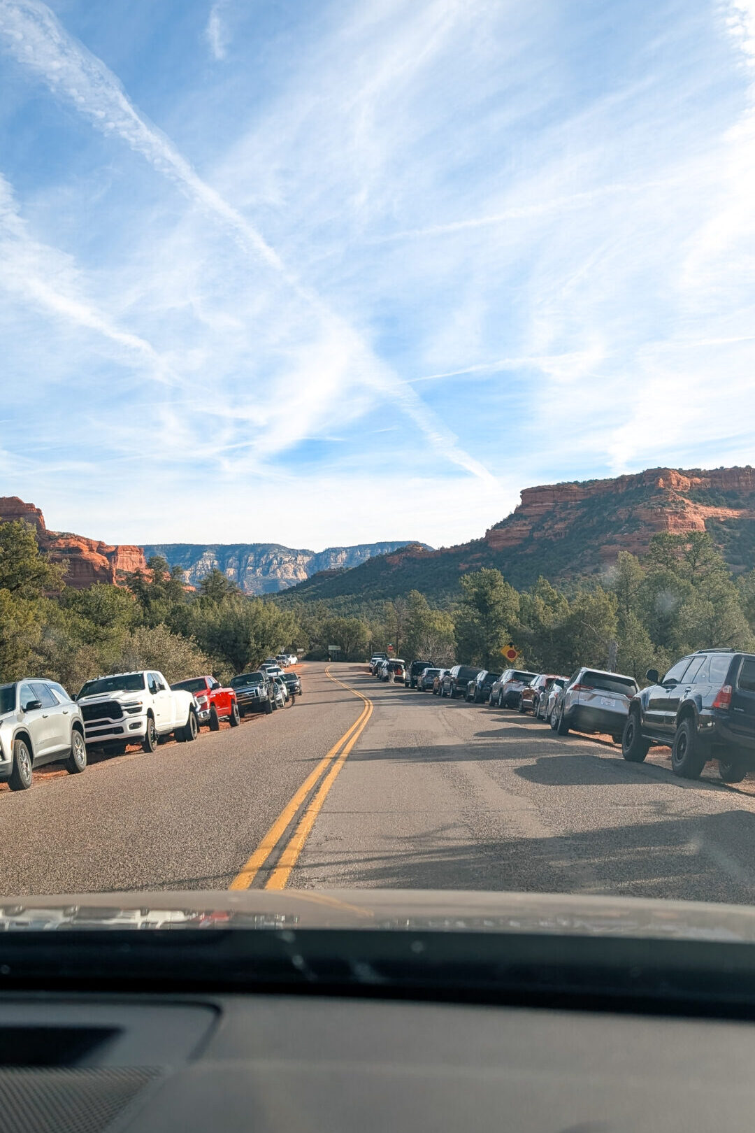 View from a windshield of a road with cars parks on both edges of the road. Red rock mountains in the distance.