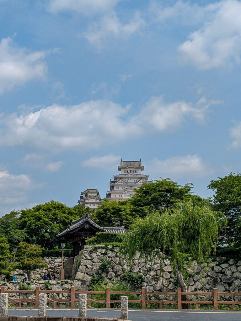 Himeji Castle in the distance, an intricate white Japanese style wood castle on top of a hill with stone walls and green trees in the foreground