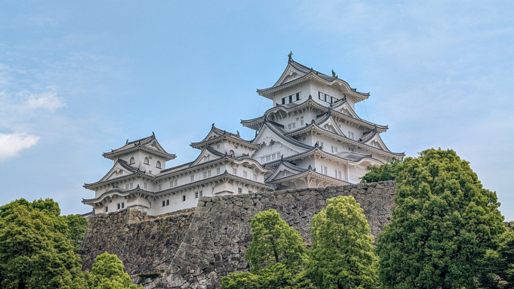 Himeji Castle, an intricate white Japanese style wood castle on top of a hill with stone walls and green trees in the foreground