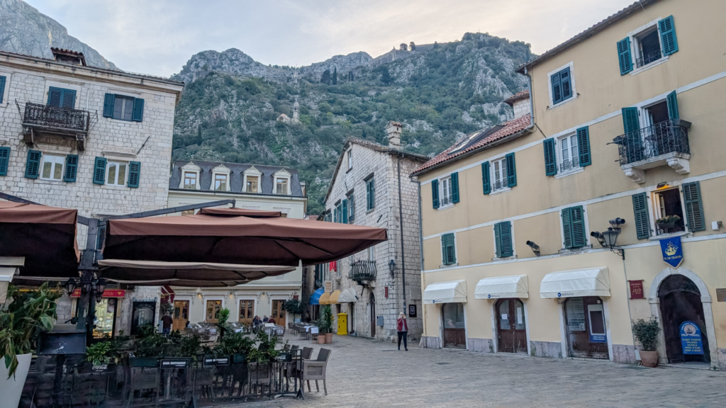 Historic stone buildings with green shutters and outdoor cafe awnings in a quiet square inside Kotor's Old Town beneath steep limestone mountains.