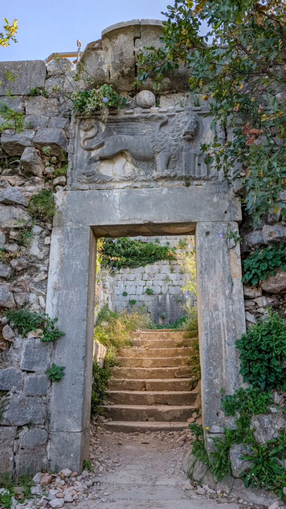 PXL_20241025_075923438.jpg:
Ancient stone archway carved with the Venetian Lion of Saint Mark, leading to stone steps on the Kotor fortress trail.