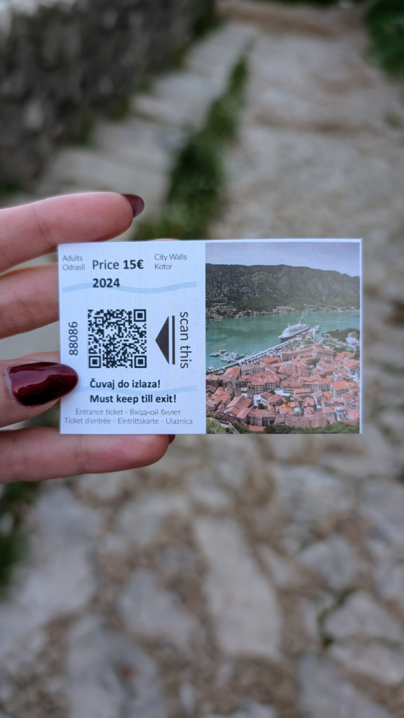 A hand holding an official 15-euro entrance ticket for the Kotor City Walls, with the trail visible in the blurred background.