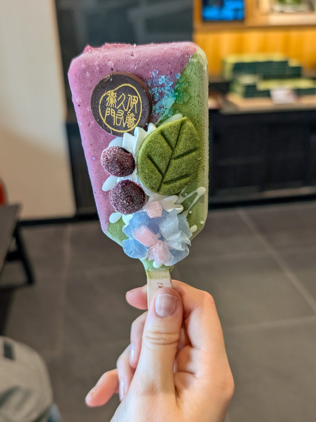 A hand holding a gourmet Japanese ice cream bar decorated with matcha tea leaves, purple glaze, and sugar flowers.