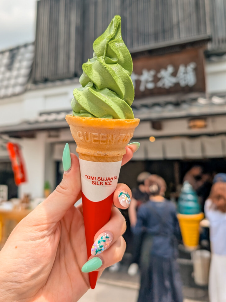 A hand holding a swirl of rich matcha soft-serve ice cream in front of a traditional tea shop in Uji.