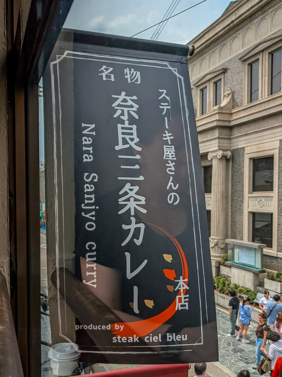 Black hanging sign for Nara Sanjo Curry seen from a window, overlooking a busy shopping street in Nara, Japan.