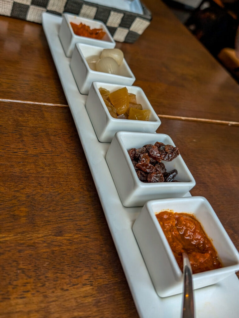 A tray of five small white square bowls containing various Japanese curry toppings, pickles, raisins, and spicy sauce on a wooden restaurant table.