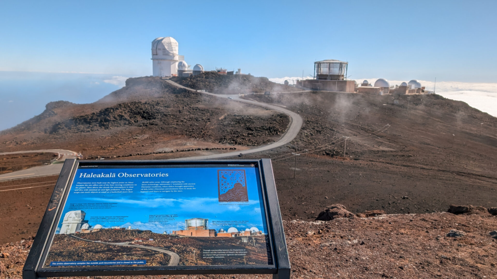 A National Park educational sign titled "Haleakalā Observatories" overlooks the rocky volcanic summit of Haleakala under a clear blue sky, with a winding access road leading to white observatory domes in the background.