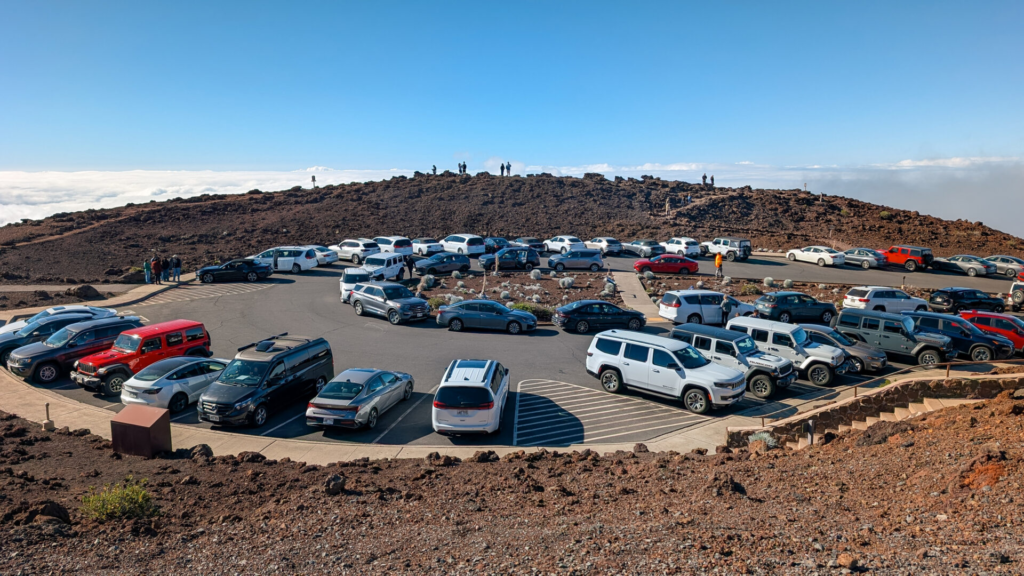 An overhead view of a packed, circular asphalt parking lot at the Haleakala summit filled with rental cars and SUVs, surrounded by red volcanic rock and a crowd of tourists gathering on a ridge above a layer of clouds.