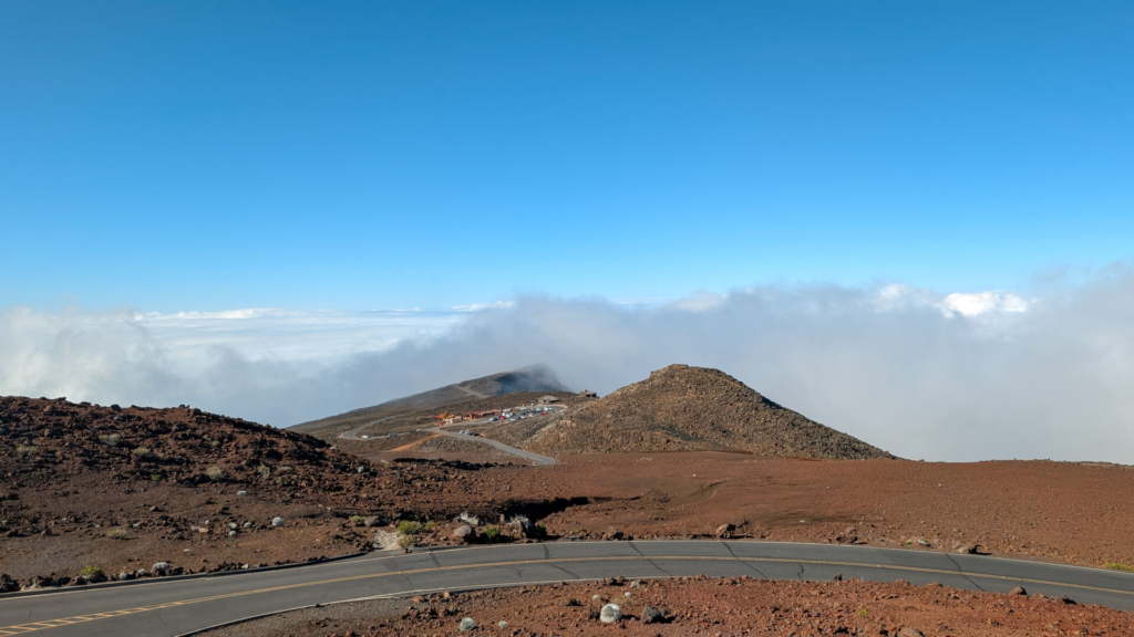 A scenic view from a winding mountain road on Haleakala showing volcanic cinder cones cutting through a thick, dramatic bank of white rolling clouds under a bright blue sky.