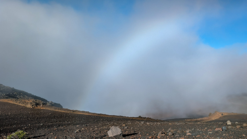 A faint white fogbow arches through a thick blanket of dense clouds and mist rolling over the dark, rocky volcanic landscape of Haleakala crater.