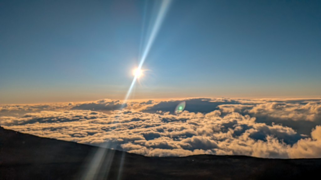 A breathtaking view from above the clouds at Haleakalā summit during the late afternoon golden hour, with the bright sun casting a dramatic, long vertical lens flare over a vast sea of rolling white clouds.
