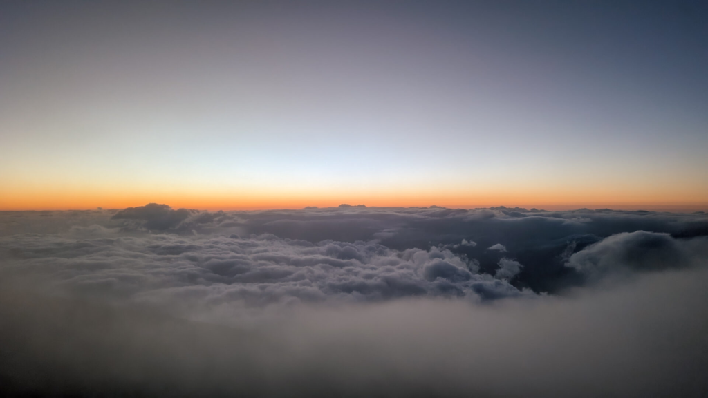 A breathtaking panoramic view after sunset at Haleakala, showing a vast, dark sea of clouds beneath a gradient sky transitioning from fiery orange at the horizon to deep blue above.