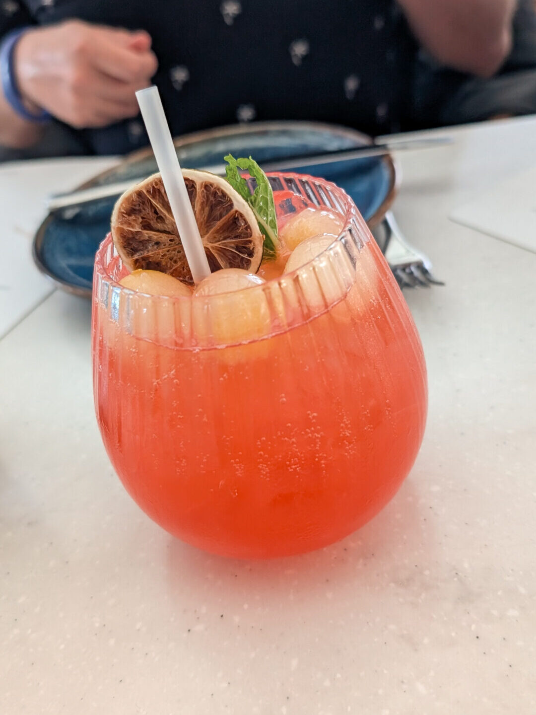 A bright coral-pink cocktail in a ribbed glass topped with melon ice spheres, a sprig of fresh mint, a dehydrated citrus wheel, and a white straw.
