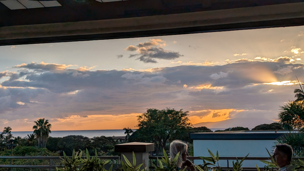 Golden sunset view overlooking the Pacific Ocean and lush palms through open-air dining patio windows, with sun rays breaking through soft clouds.