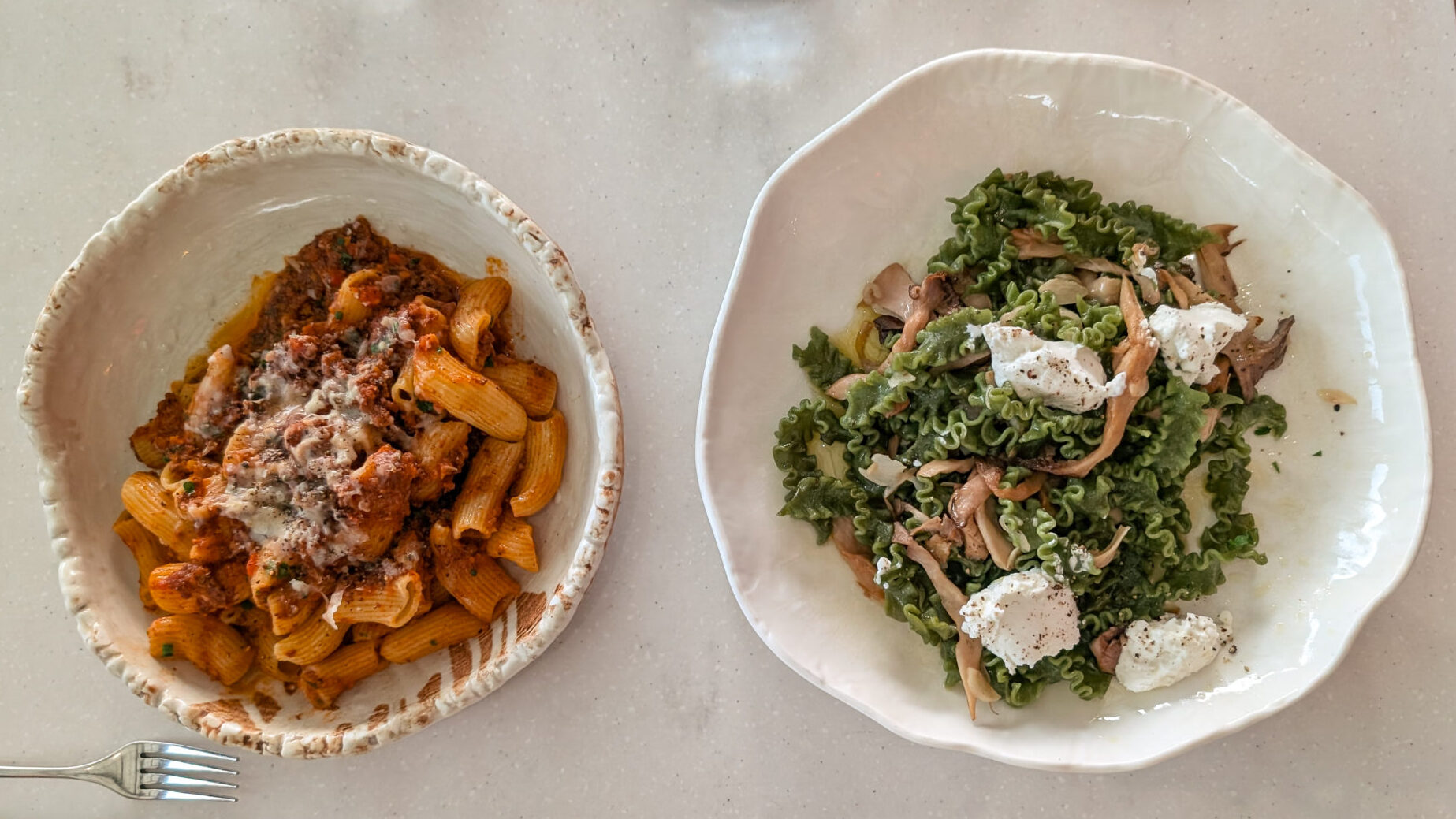 Two ceramic pasta bowls on a dining table: rigatoni in a rich meat ragù topped with melted cheese, and frilly spinach pasta tossed with wild mushrooms and dollops of local goat cheese.