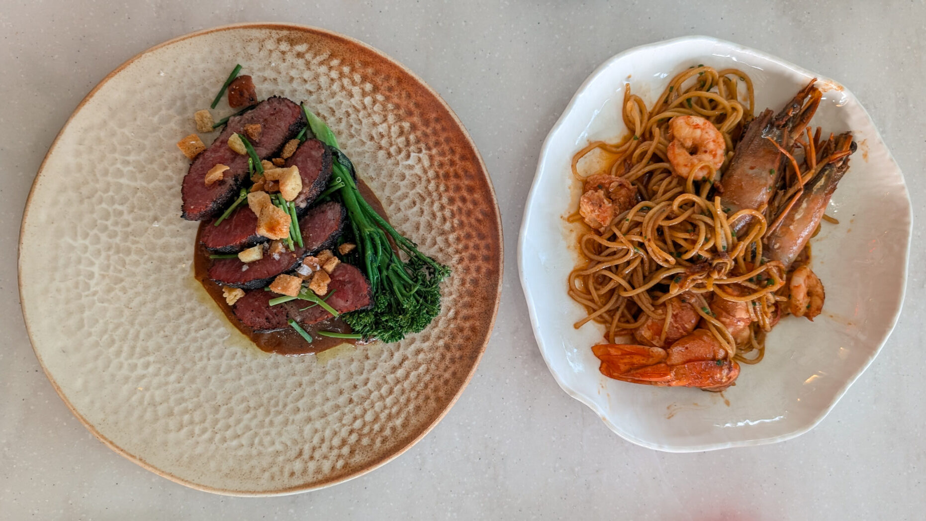 A plate of spaghettini, tossed with whole prawns and shellfish next to a textured beige plate of sliced rare steak topped with crispy garlic, chives, and tender broccolini.